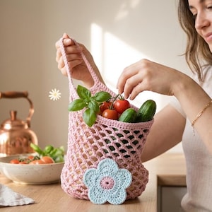 May include: A light pink crocheted produce bag filled with fresh tomatoes, cucumbers, and basil. A light blue and pink crocheted flower is placed on the wooden surface. The bag is held by a person.