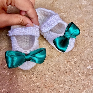 Op de afbeelding: Een paar witte gehaakte babyschoentjes met smaragdgroene satijnen strikken. De schoenen hebben een Mary Jane-stijl met een bandje over de bovenkant. De schoenen zijn handgemaakt en zien er zacht en delicaat uit.