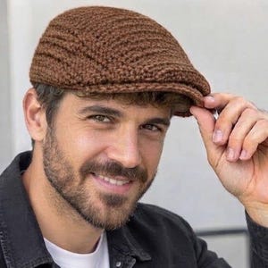 May include: A brown crocheted flat cap featuring a textured, ribbed stitch pattern. The cap has a rounded crown and a short, stiffened brim at the front, constructed from yarn with a consistent dark brown color throughout the entire piece.