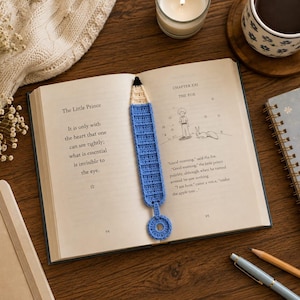 May include: A blue and cream crochet bookmark shaped like a pencil with a small circular loop at the bottom. The bookmark is placed inside an open copy of The Little Prince, featuring text and a line drawing of a boy and a fox on the pages.