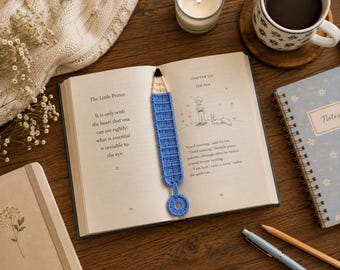 May include: A blue and cream crochet bookmark shaped like a pencil with a small circular loop at the bottom. The bookmark is placed inside an open copy of The Little Prince, featuring text and a line drawing of a boy and a fox on the pages.