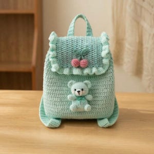 May include: A light teal crochet backpack with a handle and ruffled edges. The backpack features a cherry design and a small teddy bear applique. The bag is made of a soft, textured yarn and sits on a wooden surface.