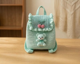 May include: A light teal crochet backpack with a handle and ruffled edges. The backpack features a cherry design and a small teddy bear applique. The bag is made of a soft, textured yarn and sits on a wooden surface.
