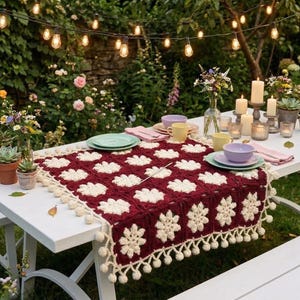 Könnte beinhalten: Ein weißer Picknicktisch ist für ein Essen im Freien gedeckt, geschmückt mit einem bordeauxroten und cremefarbenen Häkeltischläufer. Der Tisch ist mit Tellern, Tassen, Kerzen und Topfpflanzen dekoriert, mit Lichterketten darüber.
