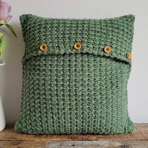 May include: A square, sage green knitted cushion with a textured pattern. The cushion features a flap secured by five round, light brown buttons. The cushion is displayed on a wooden surface, with a white vase of purple tulips in the background.