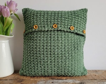 May include: A square, sage green knitted pillow with a textured pattern. The pillow features a flap secured by five round, light brown buttons. The pillow is displayed on a wooden surface, with a white vase of purple tulips in the background.