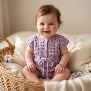 May include: A smiling baby in a lavender crochet romper with button fastenings and a tie waist sits in a wicker basket. The basket is lined with a cream blanket and pillows.