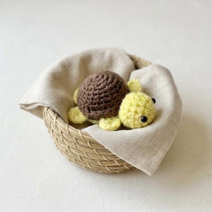 May include: A crocheted turtle, featuring a brown shell and yellow body, sits in a woven basket lined with beige fabric. The turtle has black eyes. A charming, whimsical creation.