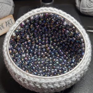 Peut inclure: Une pièce d'art géode artisanale, de forme ronde. L'intérieur est rempli de perles irisées, encadré dans un cadre en crochet gris avec un bord en métal argenté. Le signe en bois indique "MY GEODE ART".