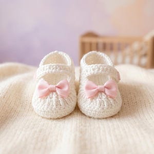 Op de afbeelding: Een paar witte gehaakte babyschoentjes met roze strikjes. Ze liggen op een cr&egrave;mekleurige gebreide deken. Op de achtergrond is een wieg te zien. Een schattig cadeau voor een pasgeboren baby.