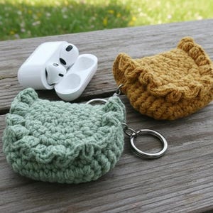 May include: A pair of white wireless earbuds next to two handmade crochet earbud cases. One case is light green, the other is mustard yellow. Both cases have a scalloped edge and a silver key ring.