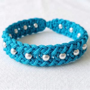 May include: A turquoise macrame bracelet with silver-colored beads. The bracelet is made of woven turquoise cord and features evenly spaced, small, round silver beads. The bracelet is displayed on a white surface.