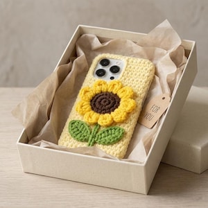 Könnte beinhalten: Eine gelbe gehäkelte Handyhülle mit Sonnenblumenmuster, präsentiert in einer Geschenkbox. Die Sonnenblume hat gelbe Blütenblätter und eine braune Mitte mit grünen Blättern. Ein kleines Etikett trägt die Aufschrift "FOR YOU".