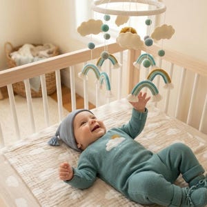 Op de afbeelding: Een baby in een blauwe outfit en muts ligt in een houten wieg en reikt naar een hangende mobiel. De mobiel heeft vilten wolken, blauwe bollen en regenboogvormige decoraties. De outfit van de baby heeft een wolk- en zonontwerp.