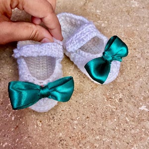 Op de afbeelding: Een paar witte gehaakte babyschoentjes met smaragdgroene satijnen strikken. De schoenen hebben een Mary Jane-stijl met een bandje over de bovenkant. De schoenen zijn handgemaakt en zien er zacht en delicaat uit.