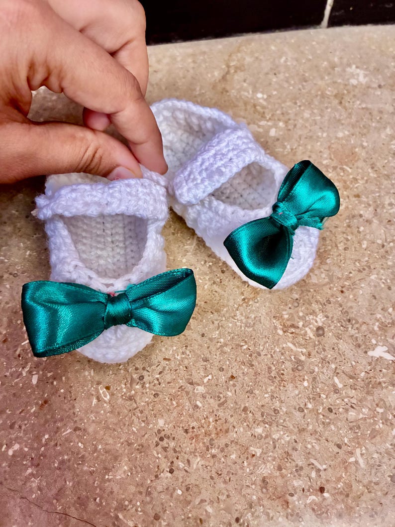 Op de afbeelding: Een paar witte gehaakte babyschoentjes met smaragdgroene satijnen strikken. De schoenen hebben een Mary Jane-stijl met een bandje over de bovenkant. De schoenen zijn handgemaakt en zien er zacht en delicaat uit.