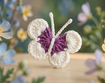 May include: A handmade crochet butterfly with white wings and a purple body. The butterfly has delicate antennae and is set against a blurred background of flowers. The craft item is a decorative piece.