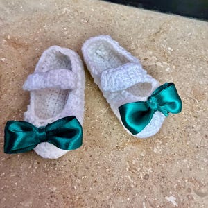 Op de afbeelding: Een paar witte gehaakte babyschoentjes met een bandje en een teal satijnen strik aan de voorkant. De handgemaakte schoentjes liggen op een lichtbruin oppervlak.