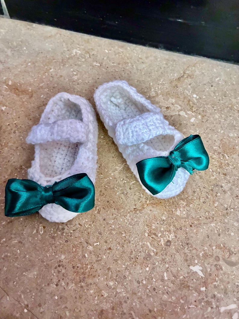 Op de afbeelding: Een paar witte gehaakte babyschoentjes met een bandje en een teal satijnen strik aan de voorkant. De handgemaakte schoentjes liggen op een lichtbruin oppervlak.