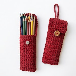Op de afbeelding: Twee handgemaakte, rode gehaakte etuis. Eén etui is open, gevuld met kleurpotloden. De andere etui is gesloten met een houten knoopsluiting. De etuis zijn gemaakt van een getextureerd, gebreid materiaal.