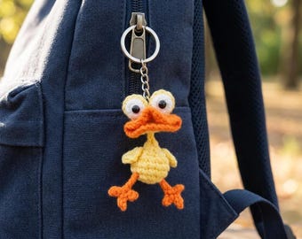 May include: A handmade yellow crochet duck keychain with orange beak, feet, and accents. The duck has large, googly eyes and is attached to a silver keychain. The keychain is hanging from a navy blue backpack zipper.