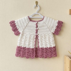 May include: A handmade, white and mauve crochet baby dress hangs on a white hanger. The dress features a scalloped hem and sleeves, with a button-up front. The background is a light beige wall.