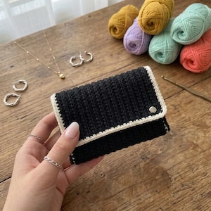 May include: A black crochet wallet with a white border and a silver snap closure. The wallet is held in a hand, with jewelry and colorful yarn balls in the background. The yarn colors include yellow, purple, green, and orange.