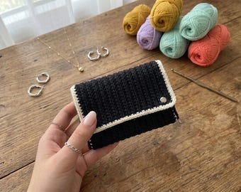 May include: A black crochet wallet with a white border and a silver snap closure. The wallet is held in a hand, with jewelry and colorful yarn balls in the background. The yarn colors include yellow, purple, green, and orange.