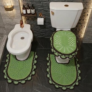May include: A bathroom set featuring a green and brown crocheted toilet seat cover and matching floor mat. The toilet and bidet are white. The set has a scalloped edge design. The bathroom has a dark tile floor and a gray stone wall.