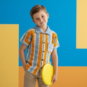 May include: A short-sleeved, button-down shirt with a crochet design in blue, yellow, and white. The shirt has a collar and a pocket with a small square design. The person is holding a yellow frisbee and wearing khaki trousers.