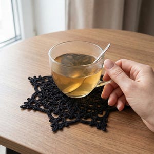 Può includere: Una tazza da tè in vetro trasparente con un cucchiaino d'argento poggia su un sottobicchiere nero all'uncinetto. La tazza contiene tè e il sottobicchiere è su un tavolo di legno.