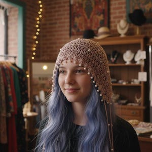 May include: A light brown crocheted hat adorned with small white beads. The hat is worn by a person with blue hair, standing in a vintage clothing store. Clothing racks and other hats are visible in the background, suggesting a bohemian or vintage fashion theme.