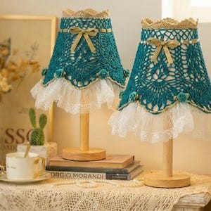 May include: Two teal crochet lamps with wooden bases and gold accents. The lampshades feature a lace trim and a bow. The lamps are on a table with books and a cup of coffee.