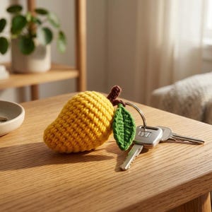 May include: A yellow crocheted mango keychain with a green leaf and brown stem, attached to a set of keys. The mango is resting on a wooden table. A small ceramic dish with keys and a potted plant are in the background.
