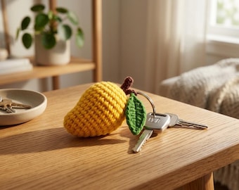 May include: A yellow crocheted mango keychain with a green leaf and brown stem, attached to a set of keys. The mango is resting on a wooden table. A small ceramic dish with keys and a potted plant are in the background.