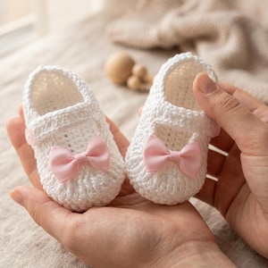 May include: A pair of handmade white crochet baby shoes with pink bows. The shoes are held in open hands, showcasing the delicate craftsmanship and soft texture. The bows add a touch of sweetness to the baby footwear.