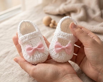 May include: A pair of handmade white crochet baby shoes with pink satin bows. The shoes are held in open hands, showcasing the delicate craftsmanship and design. The soft, textured yarn and the bows add a touch of elegance to these baby booties.