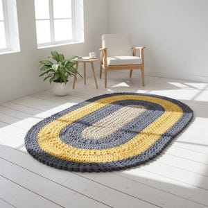 Może przedstawiać: Ręcznie robiony, owalny dywanik z szydełka o trójkolorowym wzorze. Posiada ciemnoszarą obwódkę, jasnożółtą środkową część i jasnobeżowy środek. Dywanik leży na białej drewnianej podłodze w dobrze oświetlonym pomieszczeniu.