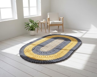 May include: An oval-shaped, handmade crochet rug with a three-tone design. The rug features a dark gray border, a bright yellow middle section, and a light beige center. The rug is placed on a white wooden floor in a well-lit room.