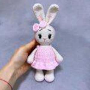 Peut inclure: Une poupée lapin au crochet faite à la main, vêtue d'une robe rose et d'un nœud assorti. Le lapin a de longues oreilles dressées, des yeux noirs et des couleurs blanches et roses. La poupée est tenue dans la main d'une personne sur un fond gris doux.
