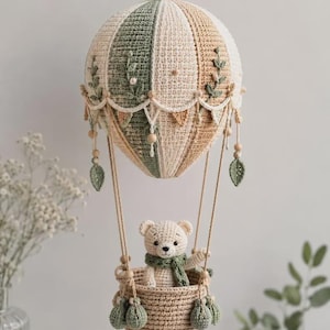 Puede incluir: Globo aerostático de ganchillo hecho a mano en crema, verde y beige. Un pequeño oso de peluche con una bufanda verde está sentado en la cesta. El globo está decorado con detalles de hojas y cuentas de madera, ideal para la decoración de la habitación de los niños.