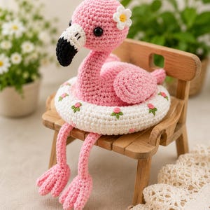 May include: A pink crocheted flamingo toy with black and white beak details and a small white flower on its head. The flamingo is sitting inside a white crocheted swim ring featuring a pattern of small pink flowers and green leaves.