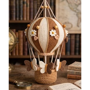 Könnte beinhalten: Handgefertigter Häkel-Heißluftballon in Braun- und Cremetönen, verziert mit weißen Gänseblümchen-Akzenten und Schmetterlingsdetails. Der Ballon ist an Schnüren aufgehängt und hat eine gewebte Korbbasis.