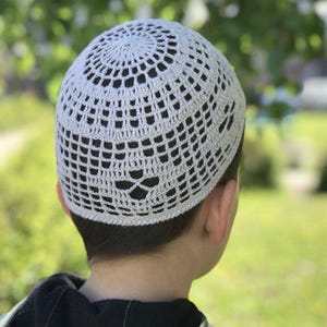 May include: A light gray crocheted skullcap with an open, lacy design. The cap is made of a soft yarn and has a circular pattern on top. The cap is worn by a person with dark hair, and the background is blurred green foliage.