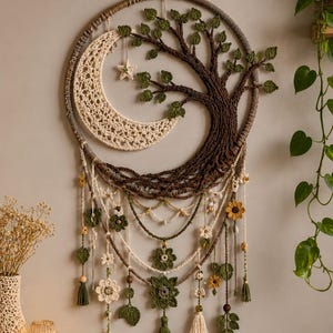 May include: A circular wall hanging featuring a crocheted crescent moon and a dark brown tree with green leaves. The piece includes hanging strands decorated with small crocheted flowers, leaves, and tassels in shades of cream, green, and yellow.