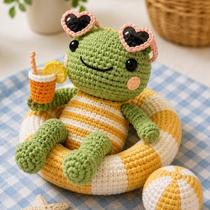 May include: A green crocheted frog toy wearing pink heart-shaped sunglasses, sitting in a yellow and white striped inflatable ring. The frog holds a small orange drink with a lemon slice and has a matching yellow and white striped ball and a starfish.