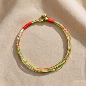 Puede incluir: Una pulsera circular construida con múltiples hilos finos multicolores en tonos rojo, verde, amarillo y blanco. Las hebras se reúnen en la parte superior en un nudo trenzado, asegurado por un envoltorio de hilo rojo sólido.