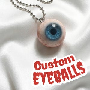 Può includere: Una collana con un ciondolo a forma di bulbo oculare realistico, con un'iride blu e una catena argentata. Il ciondolo è rotondo e dettagliato, con vene visibili. Il testo "Custom EYEBALLS" è in rosso.
