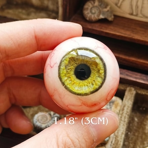 Op de afbeelding: Een realistisch protheseoog met een gele iris en zwarte pupil. Het oog heeft een diameter van ongeveer 3 cm, met zichtbare rode aderen op de witte sclera. De tekst "1.18" (3CM)" is inbegrepen.