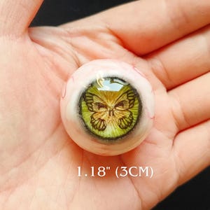 Könnte beinhalten: Ein künstliches Auge mit einem Schmetterlingsdesign. Das Auge hat einen Durchmesser von 3 cm und eine grüne Iris mit einem detaillierten Schmetterlingsmuster. Die Lederhaut ist hellrosa mit sichtbaren Adern.